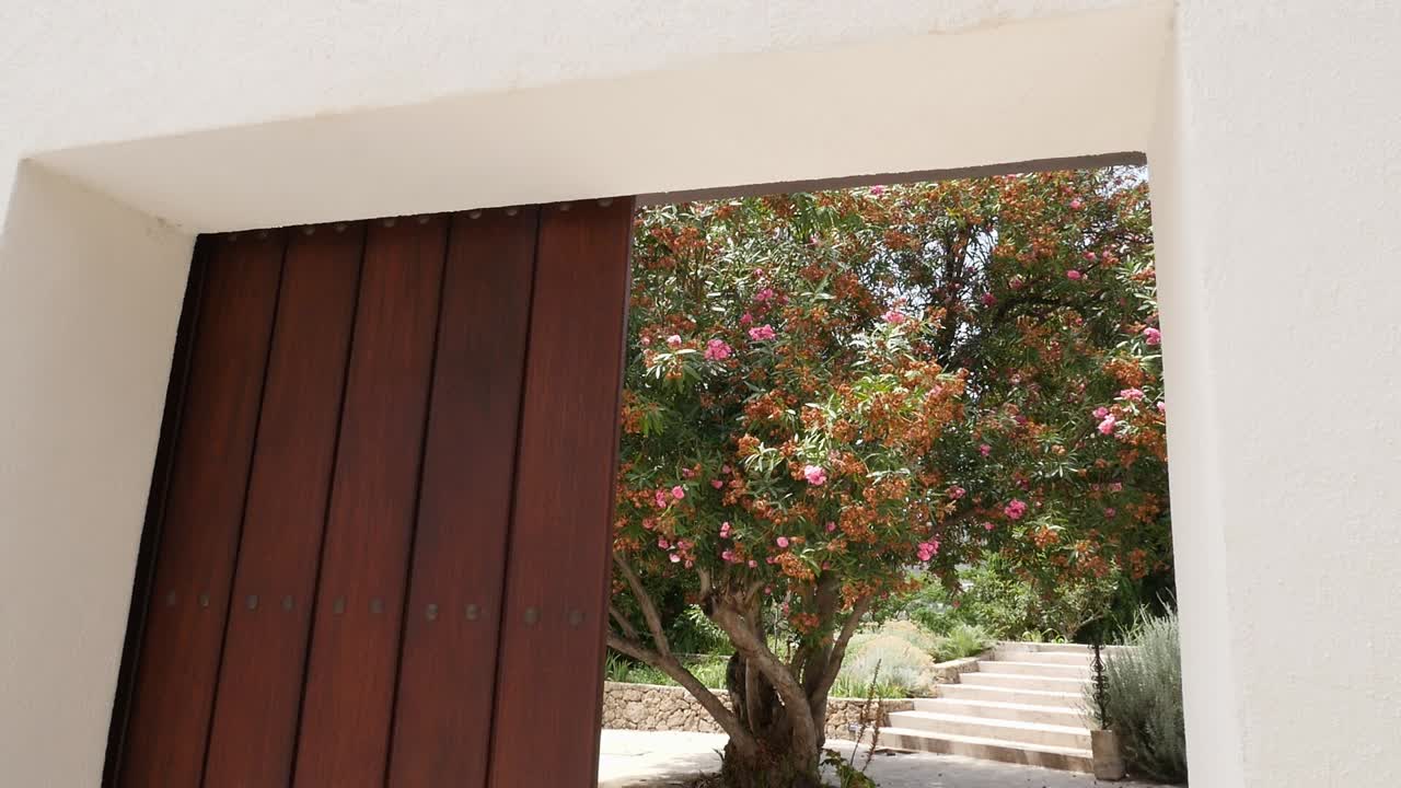 Vista da un grande cancello di legno verso un albero frondoso di oleandri in fiore e una scalinata in pietra nello storico giardino nasride di Vélez de Benaudalla, Granada