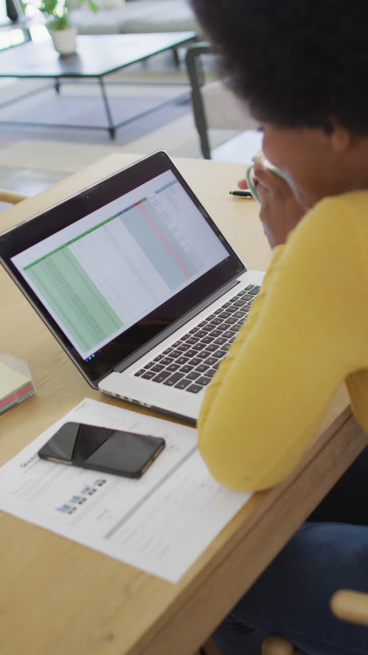 Vertical video of african american businesswoman drinking coffee and using laptop in office