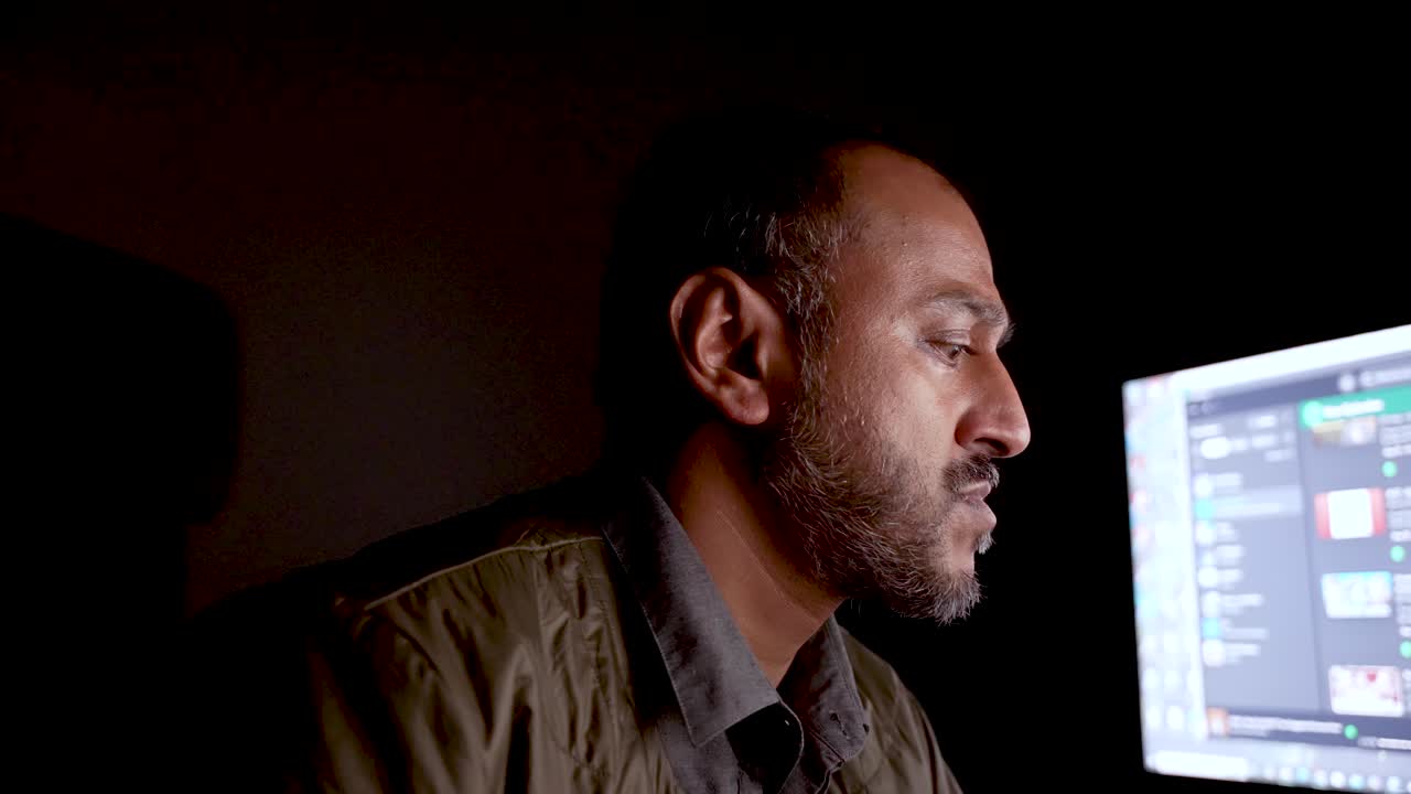 Man working diligently in a dark environment, the computer screen illuminating his face
