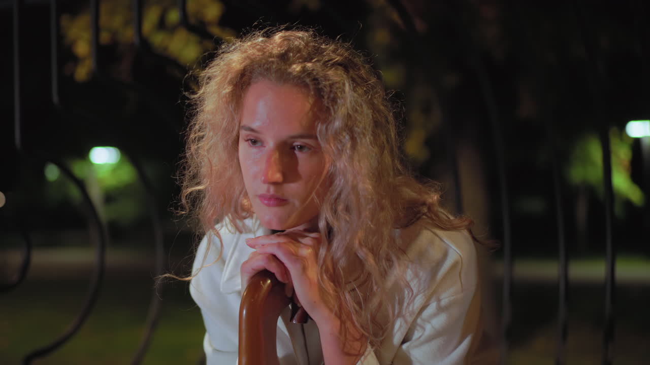 Sober woman with curly hair wearing white coat resting on umbrella handle, gazes thoughtfully into distance with soft night light creating a dreamy bokeh effect behind iron fence