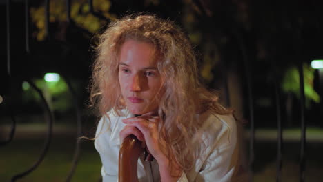sober woman with curly hair wearing white coat resting on umbrella handle, gazes thoughtfully into distance with soft night light creating a dreamy bokeh effect behind iron fence