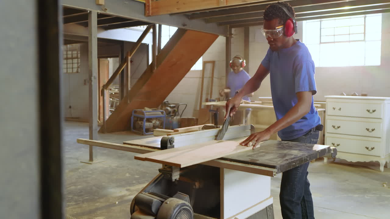 Mid Black male carpenter entering workshop cutting board on table saw while team cutting boards