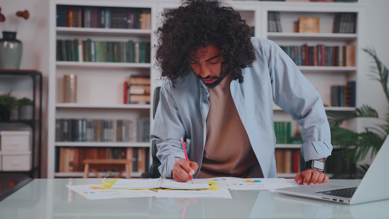 Young arabian man stand next to desk making notes looking at laptop screen