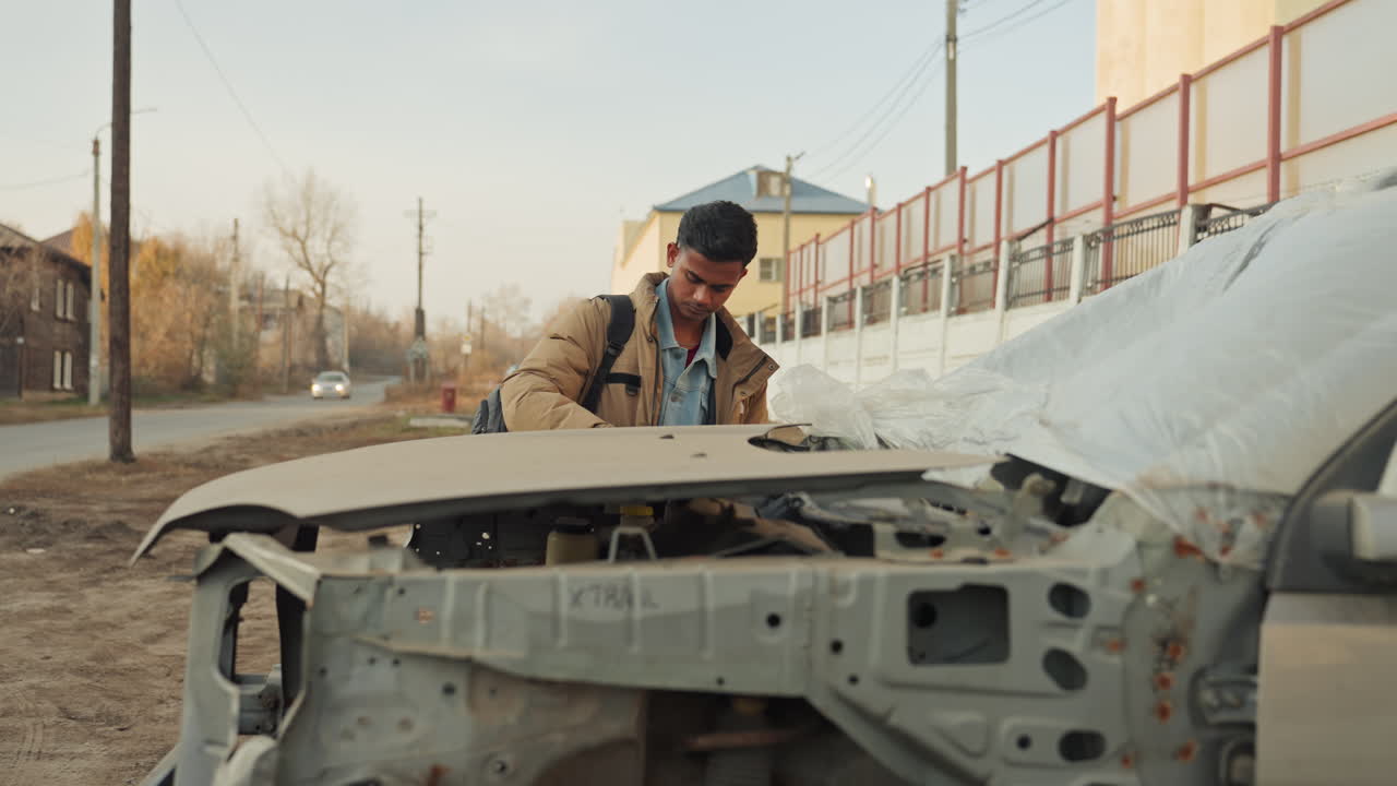 Man hammering dented front frame, hands at work, backpack nearby, muddy street scene, exposed engine bay, determined expression, striking metal with force, twilight mood, quick field repair attempt