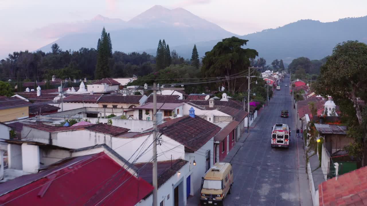 Colourful chicken bus stops to pick up people on a street corner in Antigua, Guatemala, as the sun rises with Acatenango and Fuego volcanos visible in the background and birds flying in front of drone