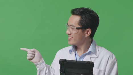 close up of asian man scientist with a tablet smiling and pointing to side while standing on the green screen background in the laboratory