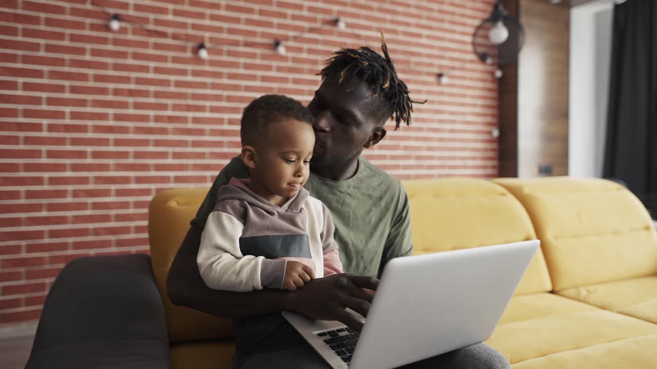 Young african american dad showing website explaining device work sit on sofa
