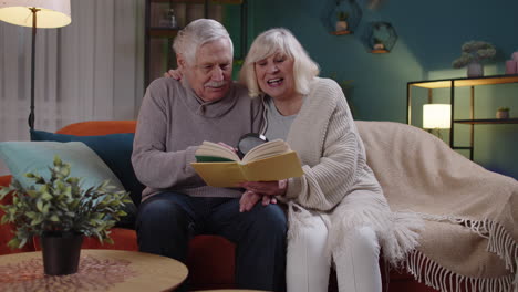 Senior-grandparents-couple-relaxing,-reading-book,-talking-enjoying-leisure-hobbies-at-night-home