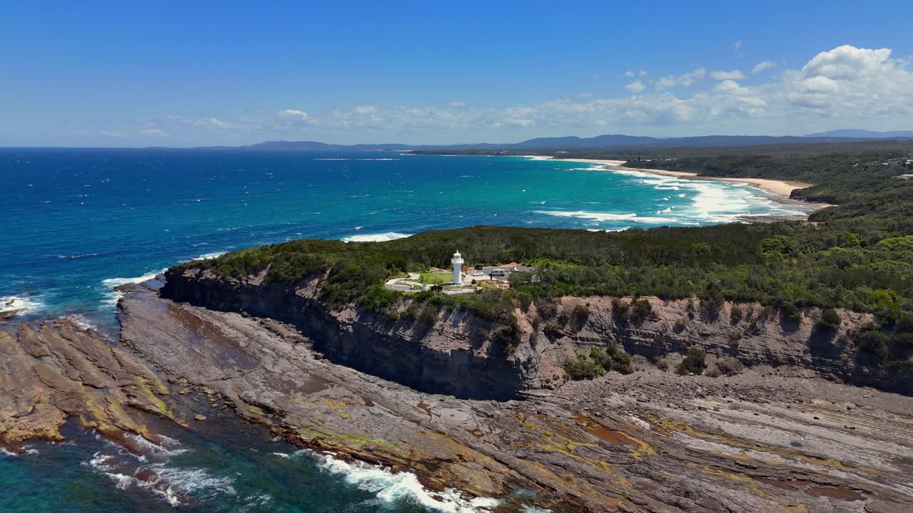 Turquoise surf hitting jagged rocks beneath cliffs of Warden Head Lighthouse point, aerial panoramic pullback