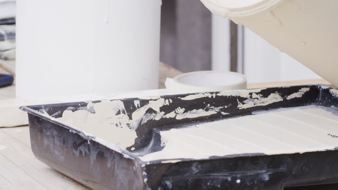 Adult painter adjusting tape pouring cream paint into tray, prepping roller for wall in room