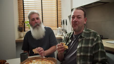 portrait of a happy lgbt couple of two men brunette in a checkered shirt and an older man with gray hair and a lush gray beard who eat pizza in the kitchen during their lunch and are happy