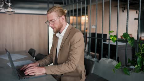 A-blond-guy-with-glasses-in-light-brown-is-at-a-laptop-and-looks-at-the-camera.-Portrait-of-a-young-male-businessman-at-his-workplace-in-a-modern-office