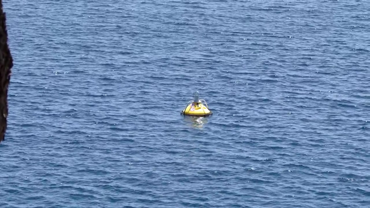 A yellow marker buoy floats on shimmering blue sea water near a rocky cliff edge.