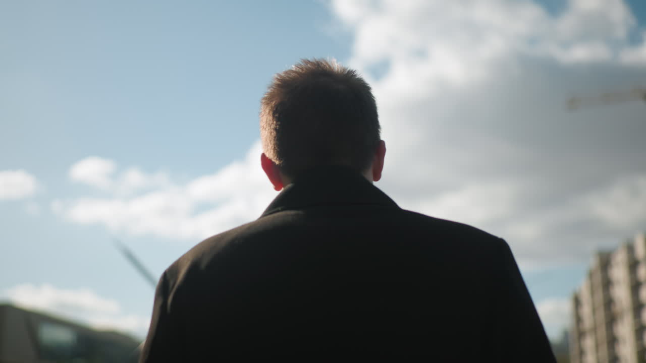 현대 도시 건축과 초록색으로 둘러싸인 구름이 있는 밝은 파란 하늘 아래에서 외부에서 자신감 있게 고 있는 검은 코트를 입은 남자의 뒷면