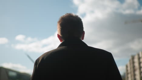 vista trasera de un hombre con abrigo negro caminando con confianza al aire libre bajo un cielo azul brillante con nubes, rodeado de arquitectura urbana moderna y vegetación
