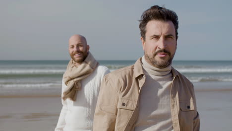 front view of a happy gay couple looking and smiling at the camera while standing on the beach