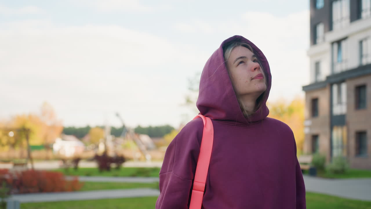 Gym enthusiast carrying workout bag walks toward iron bar gateway in outdoor fitness park framed by autumn trees pauses to tilt head and lift eyes feeling breeze teasing hoodie fabric under clear sky