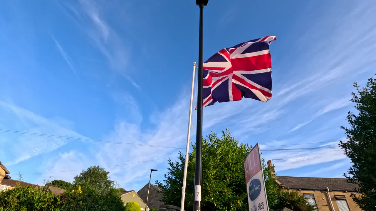 La telecamera panoramica lungo una casa in pietra e una strada, rivelando una bandiera Union Jack che sventola nella brezza sotto un cielo sereno di sera, con illuminazione naturale calda