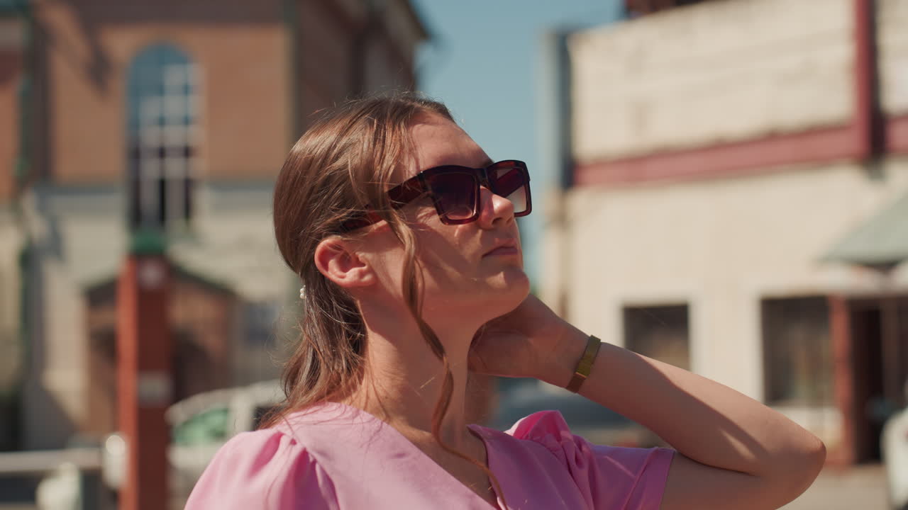 white woman in pink dress gazing toward blue sky, sunglasses perched, warm cinematic tones, brick facades and parked cars in background, relaxed neck tilt, soft smile, summer afternoon, sun flare