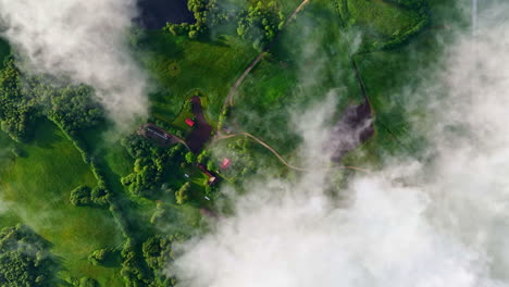an ethereal top-down drone view captures fluffy white clouds flowing over the lush latvian countryside, revealing a small village with red-roofed buildings, winding roads, and vibrant green fields.