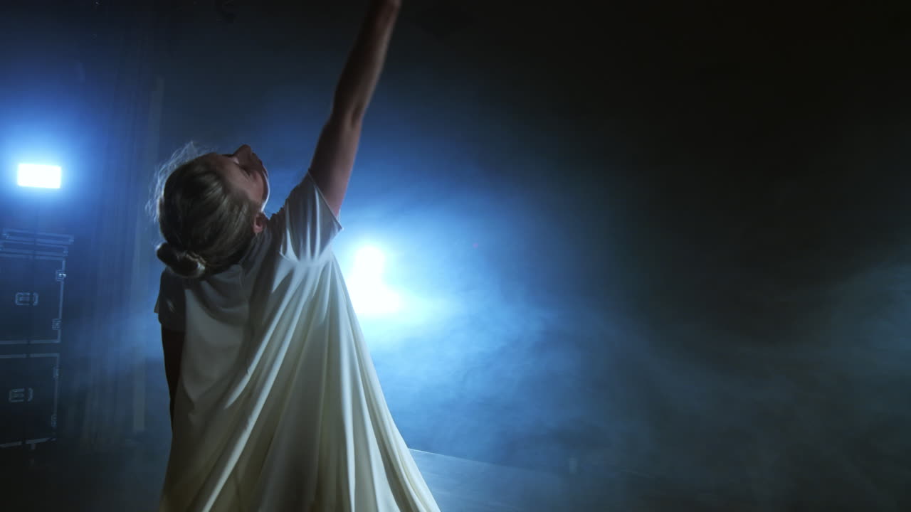 Modern dance woman in a white dress dances a modern ballet jumps makes rotation on the stage with smoke in the blue spotlights.
