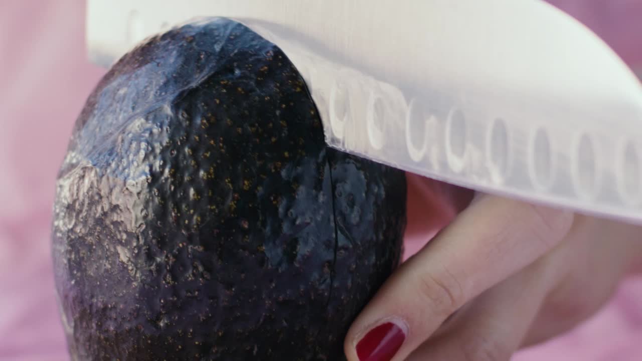 деталь авокадо, разрезаемого большим кухонным ножом, фон розовый, съемка в замедленной съемке при естественном освещении