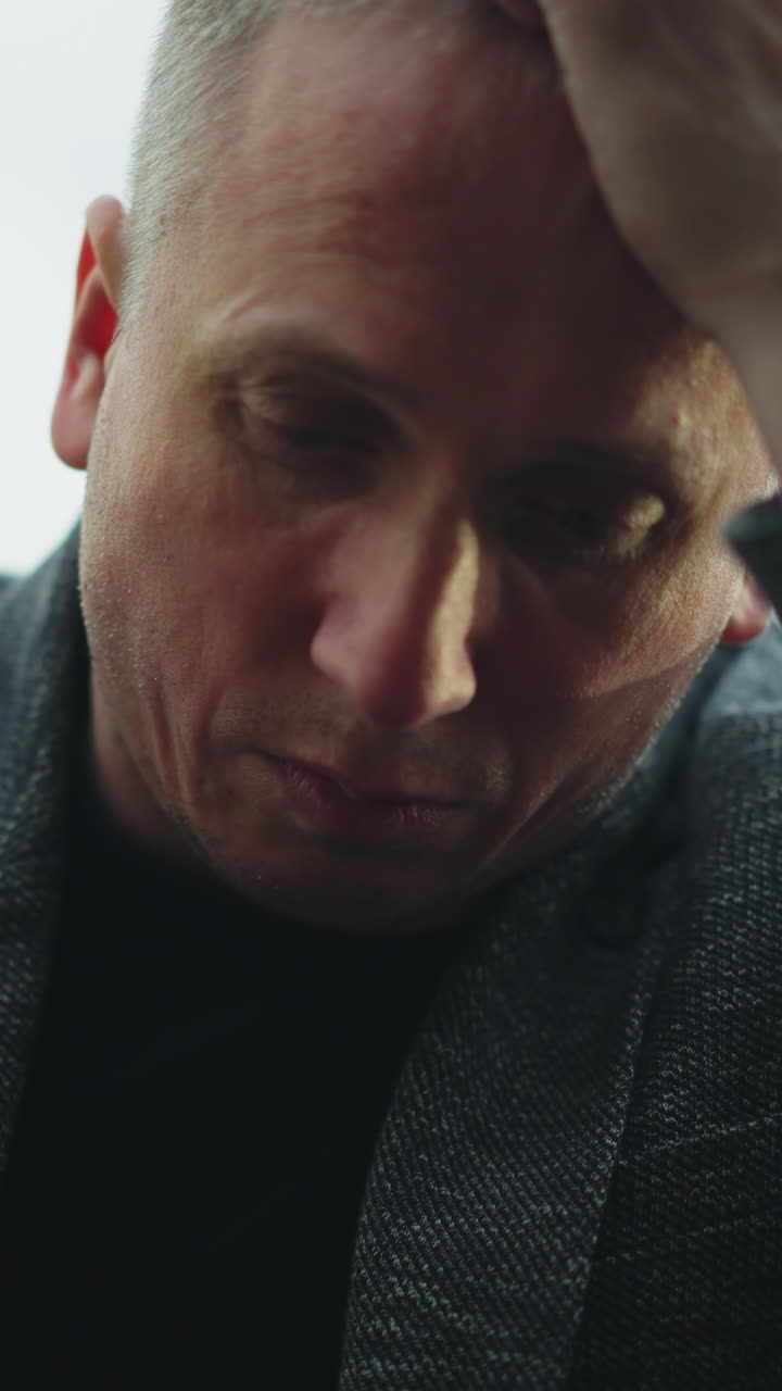 A close view of a man in a gray jacket and blue jeans sitting on a railway track, holding a handgun with his head bowed and hand resting on his head, with a blurred green forest