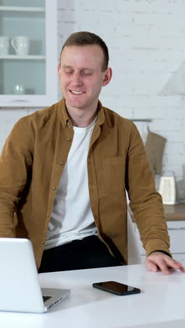 young man works and drinks tea in the kitchen. happy student studying at home during quarantine. guy using a laptop for job in comfortable apartment. vertical video
