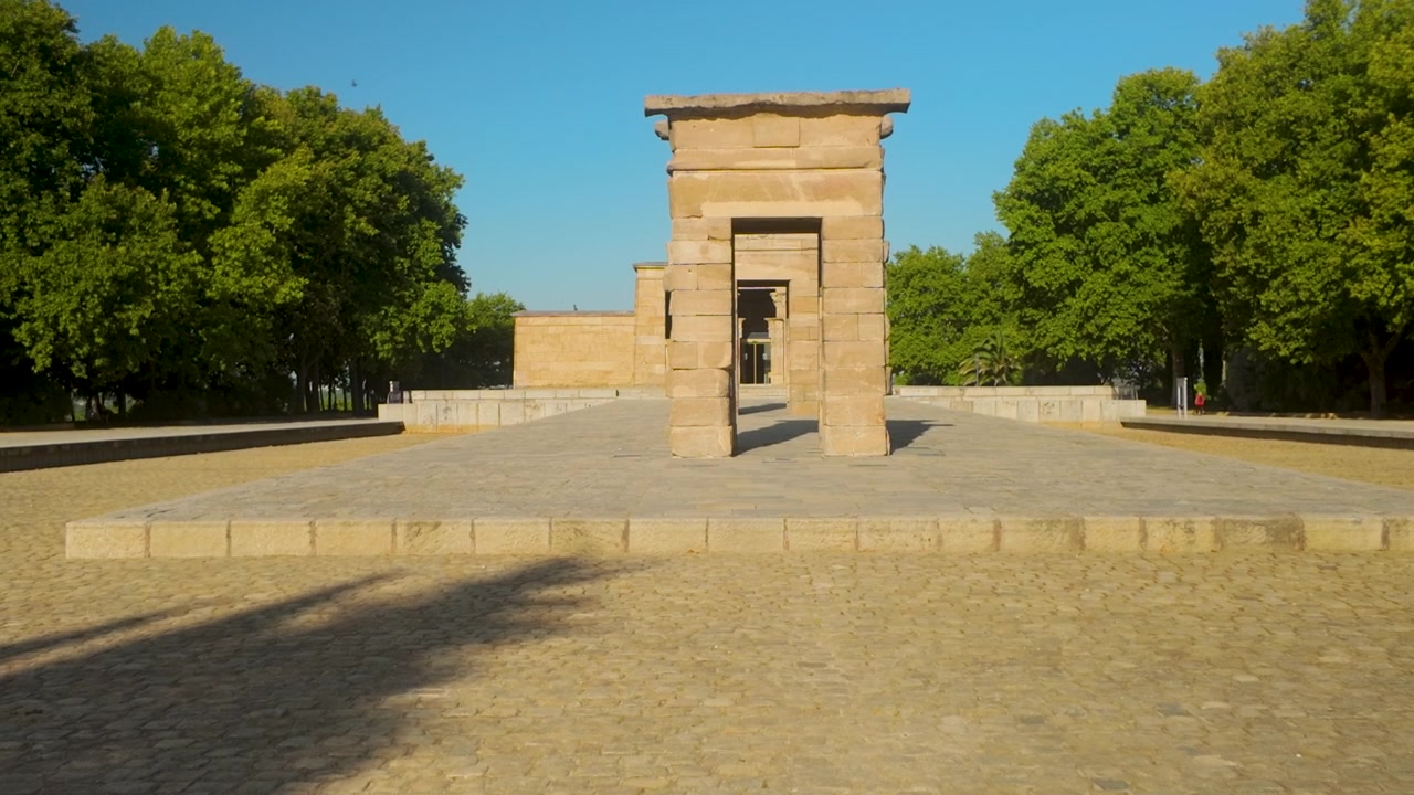 Una toma amplia del Templo de Debod, un antiguo templo egipcio reubicado en Madrid, rodeado de exuberantes árboles verdes bajo un cielo azul claro.