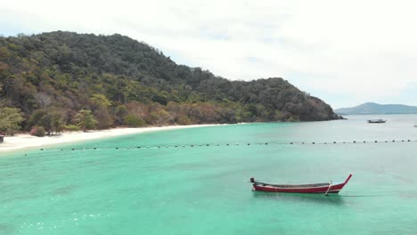 Long-tail-fishing-boat-resting-above-scenic-turquoise-shallow-sea-in-Banana-Beach-in-Koh-Hey-,-Thailand---Aerial-Low-angle-Fly-over-shot