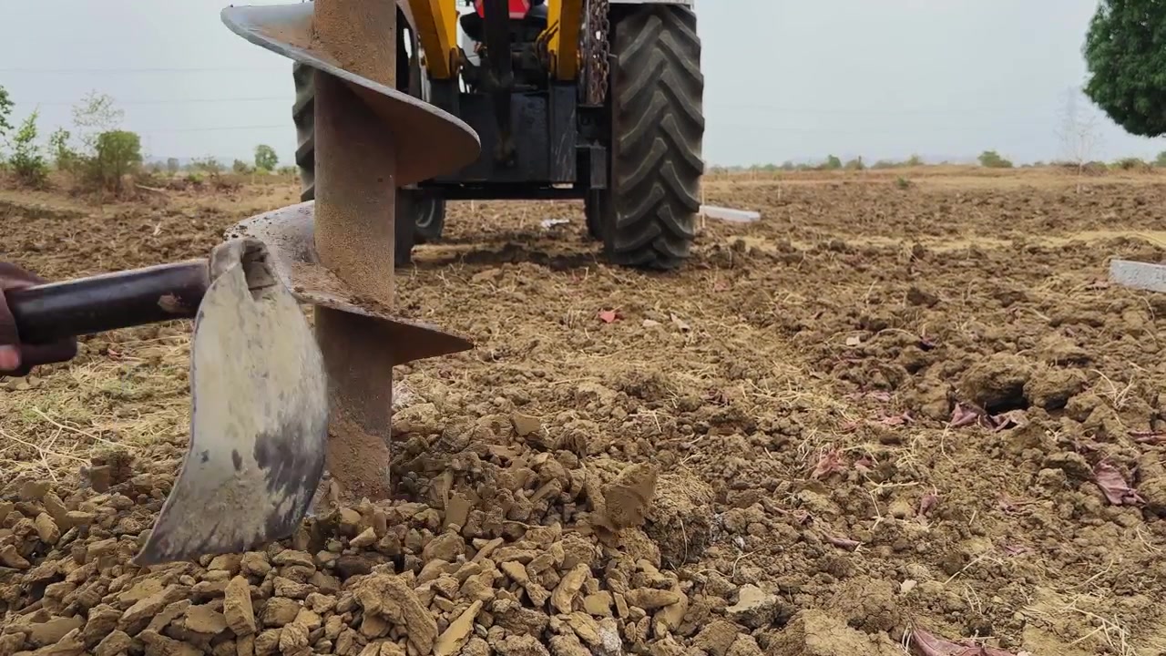 Vista aproximada de uma broca de trado posicionada no solo com um trator ao fundo e ferramenta de trabalhador visível, mostrando o processo ativo de perfuração do solo em um cenário agrícola rural