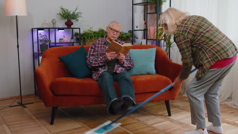 senior grandmother cleans wipes dust on floor with mop under grandfather legs while he reading book