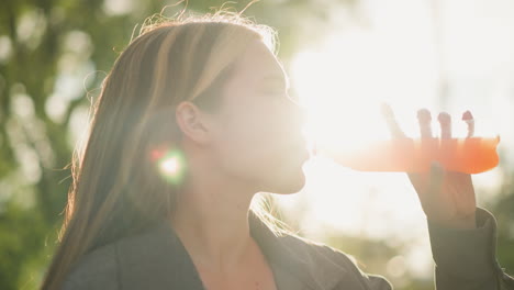 woman seated outdoors drinking orange juice from bottle with relaxed satisfaction, her expression reflecting contentment, background shows soft glow from sunlight, trees, and greenery