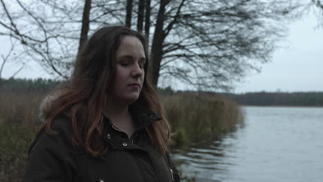 an unhappy woman standing alone at the lake and try to hold herself to think loud by touching her mouth with her hands and wipe her tear