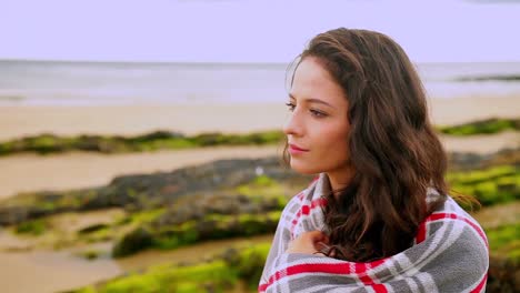 thoughtful brunette wrapped in a blanket sitting by the beach