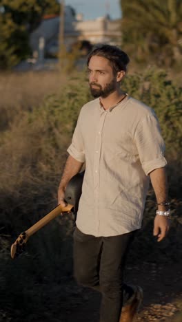 man walking through a field holding a guitar at golden hour