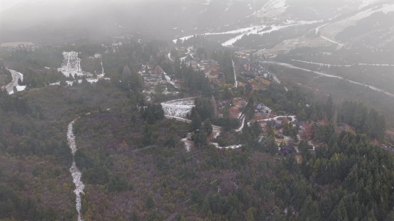 Wide aerial retreat pulls away over chalet neighborhoods and snowy ski runs on Cerro Catedral, clouds and fog enveloping the slopes above Bariloche, Patagonia