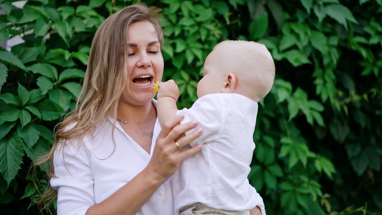Happy Caucasian woman holding her beloved baby outdoors. Mother talks to her son and he gives her a flower to smell.