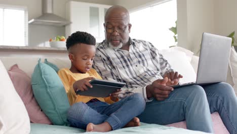 african american grandfather with grandson using tablet and laptop on couch at home, slow motion