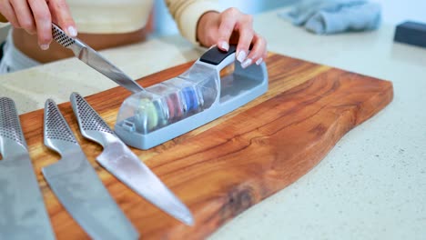 a person sharpens multiple kitchen knives using a manual whetstone sharpener on a wooden cutting board in a brightly lit kitchen