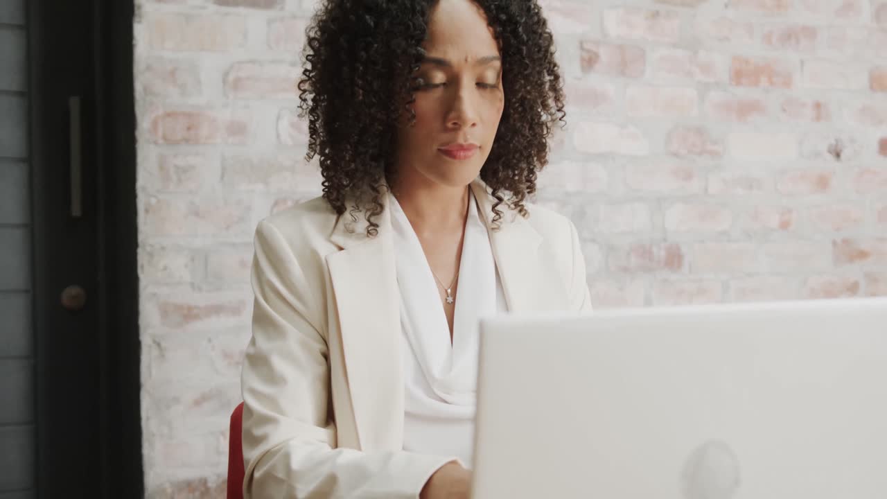 Thoughtful biracial businesswoman sitting at desk using laptop at office, in slow motion