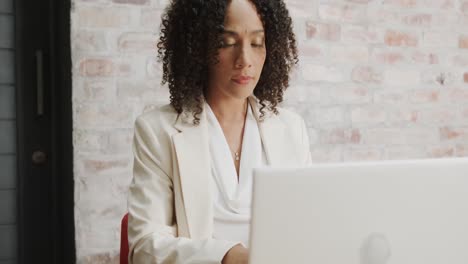 Thoughtful-biracial-businesswoman-sitting-at-desk-using-laptop-at-office,-in-slow-motion