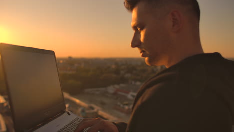 Un-Corredor-De-Bolsa-Independiente-Se-Encuentra-En-Una-Azotea-Al-Atardecer-Con-Una-Computadora-Portátil-Y-Escribe-En-Un-Teclado-Con-Los-Dedos-Mirando-El-Paisaje-Urbano-A-Vista-De-Pájaro.