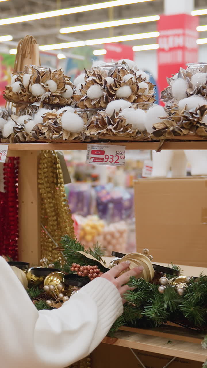 Back view of lady in white sweater touching Christmas decor in a well-lit decoration shop, shelves filled with colorful ornaments, garlands, and festive decorations