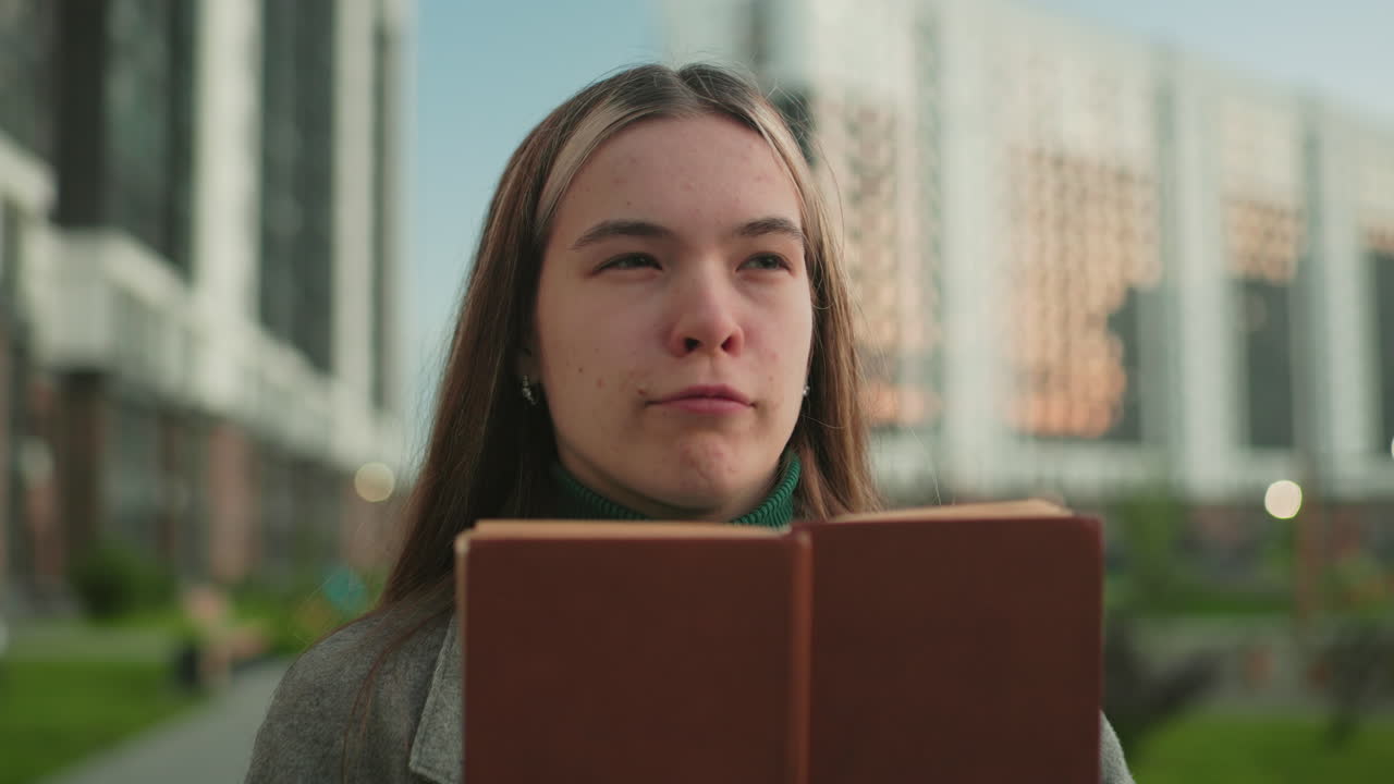 Close up of girl in gray coat playfully covering face with book then gently removing it with funny expression, blur passerby and tall building in background