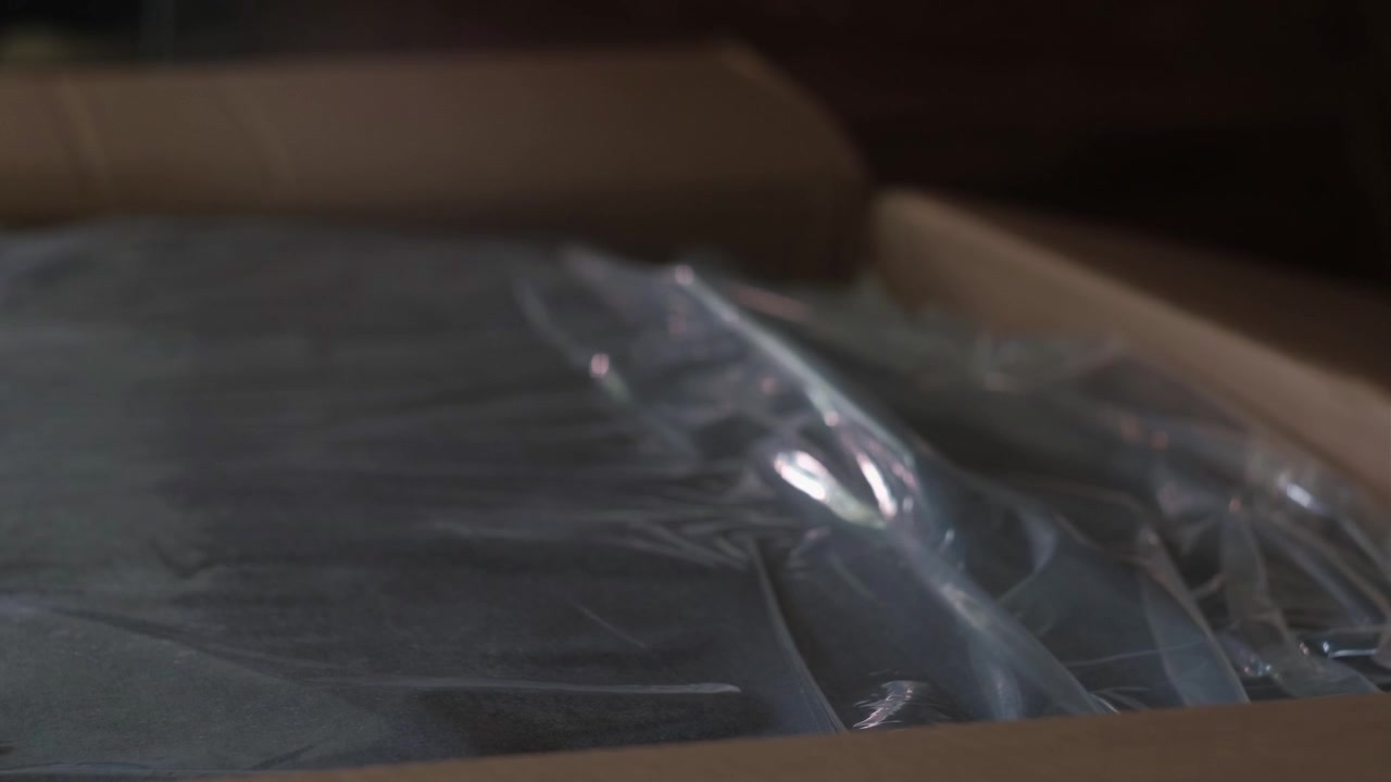 Close-up static shot of hands opening a large cardboard box indoors with visible packaging and dim lighting