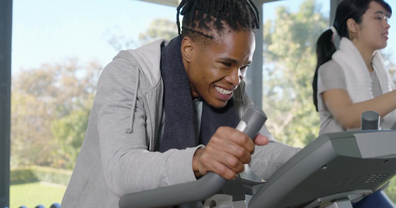 Couple exercising in bright gym with man responding to console, pushing bike, wiping towel