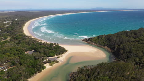 Wide-cinematic-drone-shot-of-the-South-Pacific-Ocean-and-Korogoro-Creek-with-wind-blowing-sand-across-a-sand-bar-at-Hat-Head-New-South-Wales,-Australia