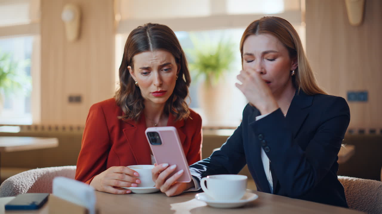 Two businesswomen frustrated cellphone message sitting at restaurant closeup