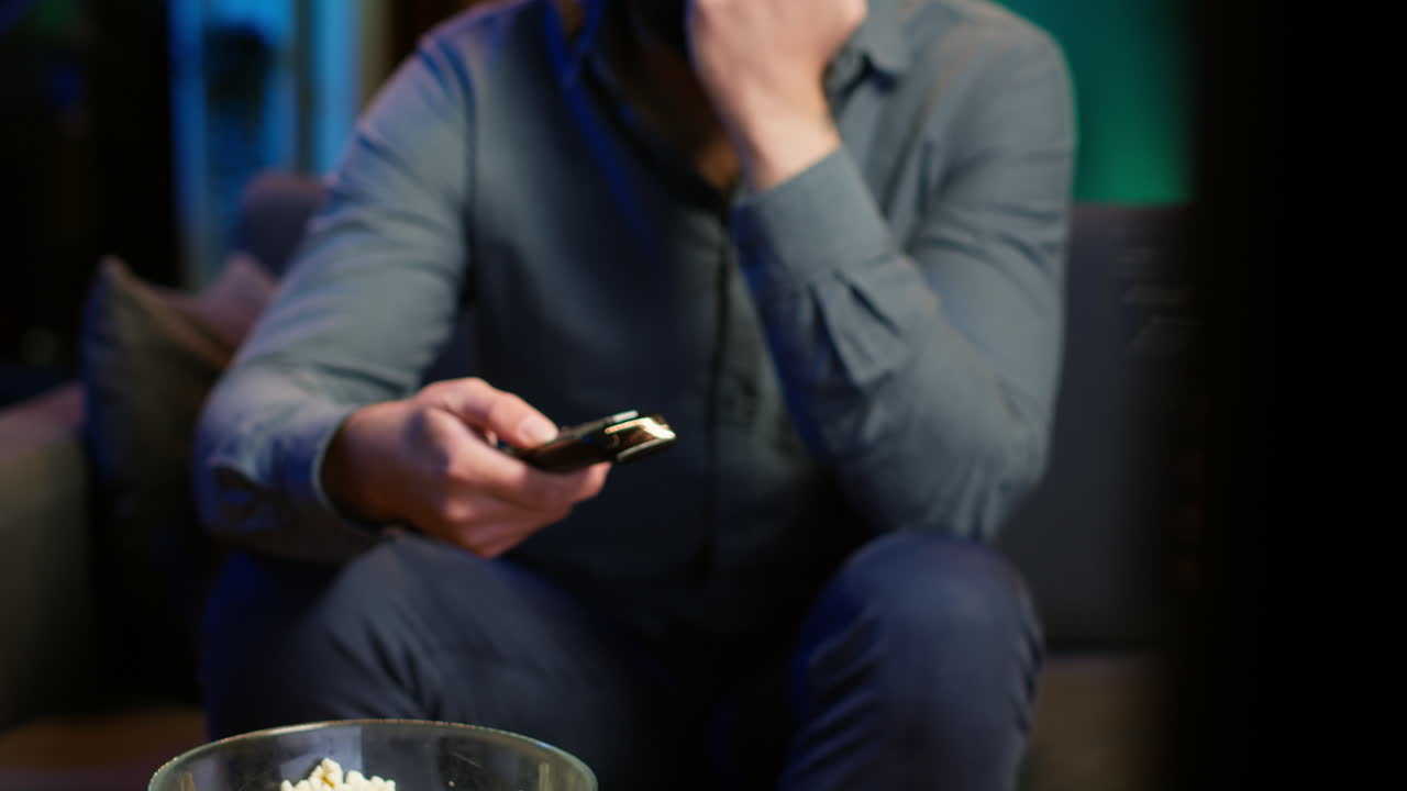 Close up of man on couch using remote control flipping through channels on TV